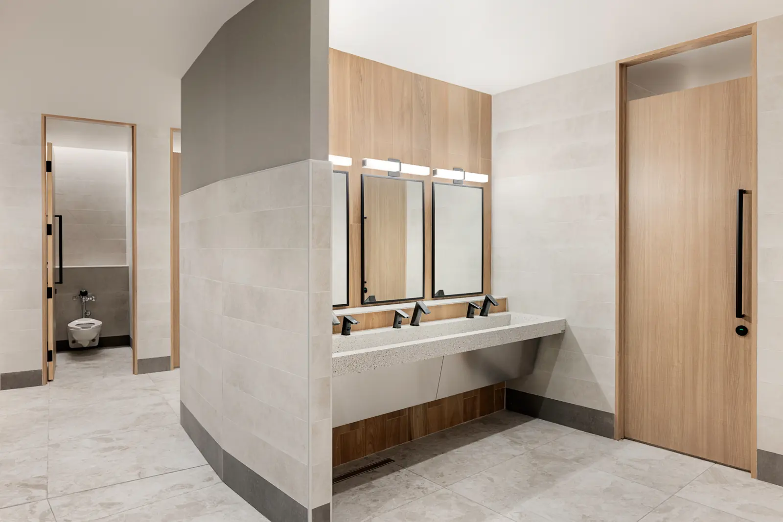 City Hall gender-neutral washroom — Finished washroom — full-height tile, wood-look privacy stalls, and a shared trough sink